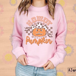 May include: A light pink sweatshirt with the text "Howdy Pumpkin" in orange and brown. The graphic features a pumpkin wearing a cow print cowboy hat, bats, and stars. The sweatshirt is a long-sleeved crew neck.
