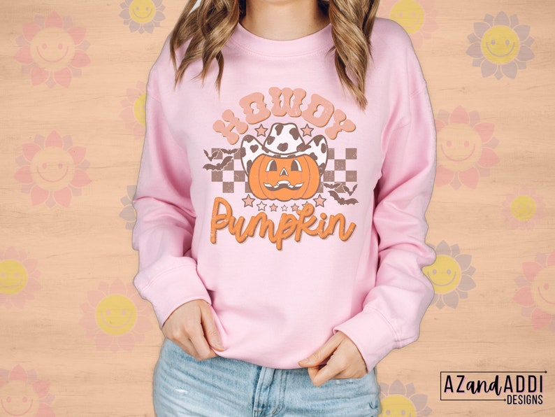 May include: A light pink sweatshirt with the text "Howdy Pumpkin" in orange and brown. The graphic features a pumpkin wearing a cow print cowboy hat, bats, and stars. The sweatshirt is a long-sleeved crew neck.