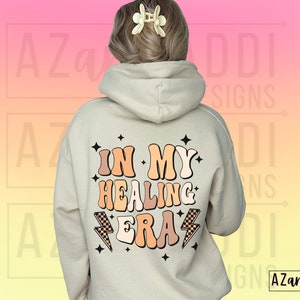 K&ouml;nnte beinhalten: Ein beiger Hoodie mit dem Text "In My Healing Era" in einer Retro-Schriftart mit Blitzen und Sternen. Der Text ist in den Farben Pfirsich, Braun und Orange gehalten.
