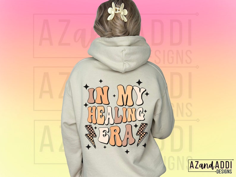 K&ouml;nnte beinhalten: Ein beiger Hoodie mit dem Text "In My Healing Era" in einer Retro-Schriftart mit Blitzen und Sternen. Der Text ist in den Farben Pfirsich, Braun und Orange gehalten.