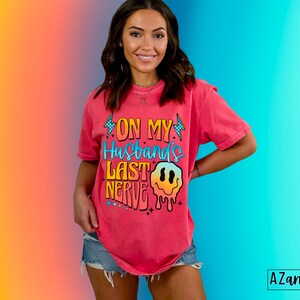 May include: A pink t-shirt with a graphic design that says "On My Husband's Last Nerve" with a dripping smiley face.