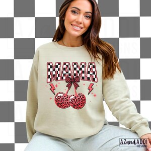 May include: A light beige sweatshirt with the word "MAMA" in black and white checkered letters. The letters are outlined in pink. Below the word "MAMA" is a pink bow with a cherry stem and two red cherries with a leopard print pattern.