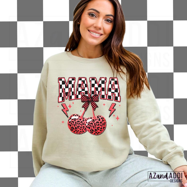 May include: A light beige sweatshirt with the word "MAMA" in black and white checkered letters. The letters are outlined in pink. Below the word "MAMA" is a pink bow with a cherry stem and two red cherries with a leopard print pattern.