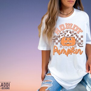 May include: White t-shirt featuring a pumpkin wearing a cow print cowboy hat. The shirt has the text "Howdy Pumpkin" in orange and peach colors. The design includes stars, bats, and a checkered pattern. The shirt is part of a fall or Halloween theme.