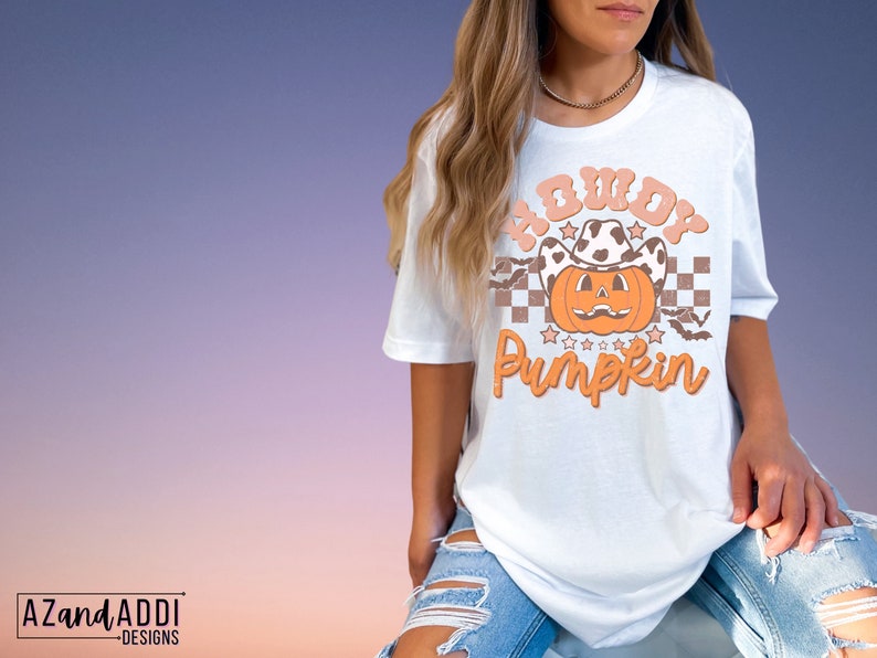 May include: White t-shirt featuring a pumpkin wearing a cow print cowboy hat. The shirt has the text "Howdy Pumpkin" in orange and peach colors. The design includes stars, bats, and a checkered pattern. The shirt is part of a fall or Halloween theme.