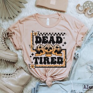 Op de afbeelding: Een roze T-shirt met een grafische print van drie schedels met luipaardprint en de tekst "Dead Tired" in zwart. De grafiek is omgeven door een zwart frame met een geruit patroon en sterren.