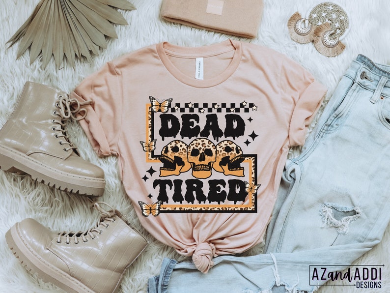 Op de afbeelding: Een roze T-shirt met een grafische print van drie schedels met luipaardprint en de tekst "Dead Tired" in zwart. De grafiek is omgeven door een zwart frame met een geruit patroon en sterren.