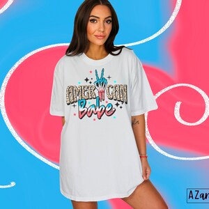 May include: White oversized t-shirt with a graphic print of a peace sign in red, white and blue colours and the text "American Babe" in a glittery font.