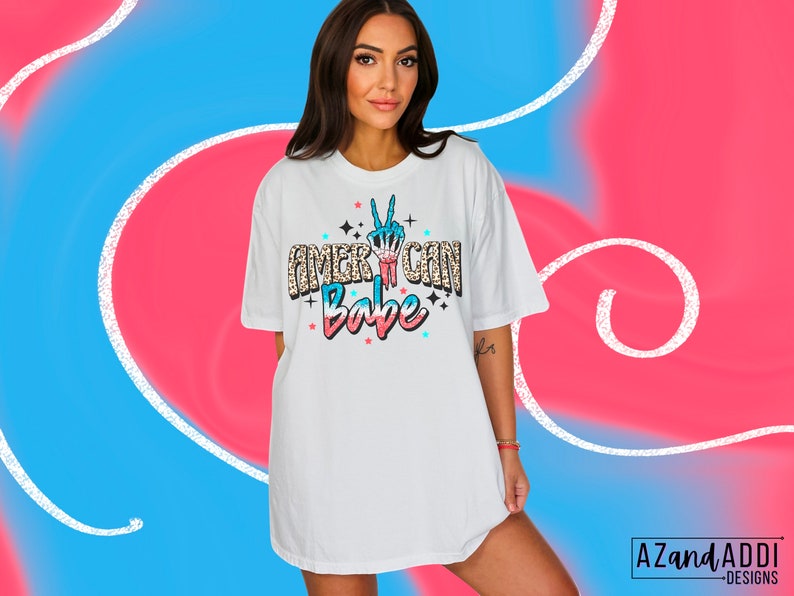 May include: White oversized t-shirt with a graphic print of a peace sign in red, white and blue colours and the text "American Babe" in a glittery font.