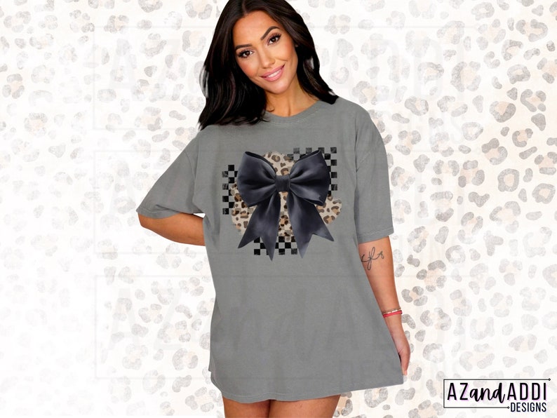 May include: A gray oversized t-shirt with a black bow on a checkered and leopard print background.