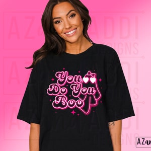 K&ouml;nnte beinhalten: Schwarzes T-Shirt mit dem Schriftzug "You Do You Boo" in einem Retro-Schriftstil, begleitet von einer Geistergrafik mit herzf&ouml;rmigen Augen und einer Schleife. Das Design ist in Pink und Wei&szlig; gehalten.