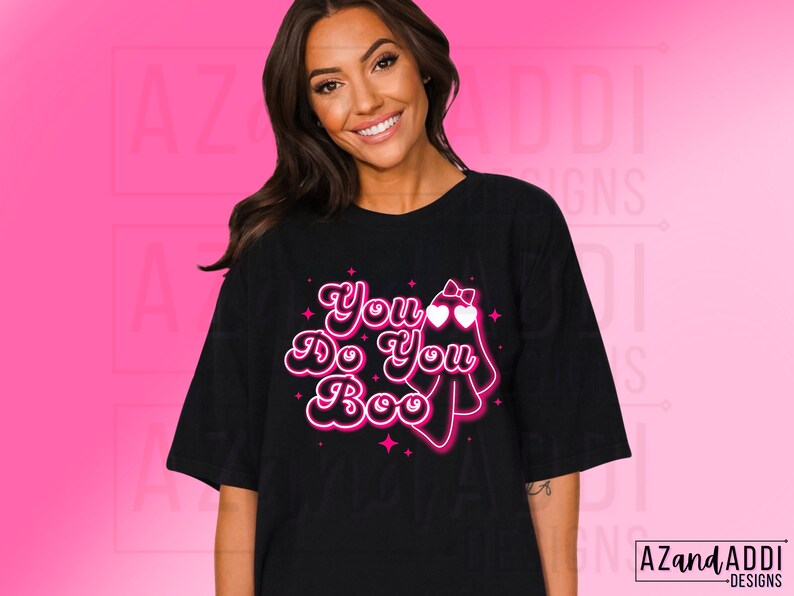 K&ouml;nnte beinhalten: Schwarzes T-Shirt mit dem Schriftzug "You Do You Boo" in einem Retro-Schriftstil, begleitet von einer Geistergrafik mit herzf&ouml;rmigen Augen und einer Schleife. Das Design ist in Pink und Wei&szlig; gehalten.