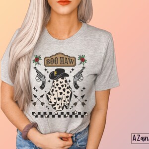 Puede incluir: Una camiseta gris con un fantasma que lleva un sombrero de vaquero y un patr&oacute;n de lunares blanco y negro. El texto "BOO HAW" est&aacute; por encima del fantasma. Tambi&eacute;n hay rosas rojas, pistolas y flechas en la camiseta.