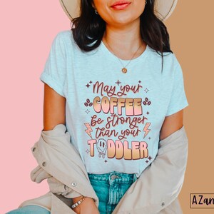 May include: A white t-shirt with a pink and orange graphic that says "May your coffee be stronger than your toddler".