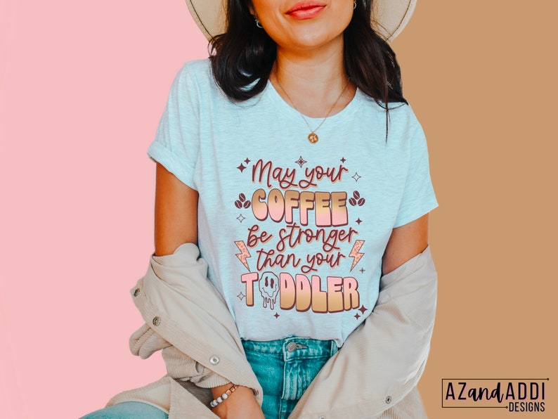 May include: A white t-shirt with a pink and orange graphic that says "May your coffee be stronger than your toddler".
