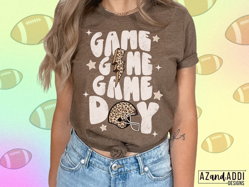 May include: A brown t-shirt with the text "Game Game Day" in white with a leopard print football helmet graphic. The shirt has a distressed look and stars around the text.