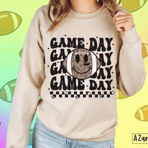 May include: A beige sweatshirt featuring the text "GAME DAY" repeated three times, with a football graphic in the middle. The football has a smiling face and a leopard print pattern. The sweatshirt has a crew neck and long sleeves.