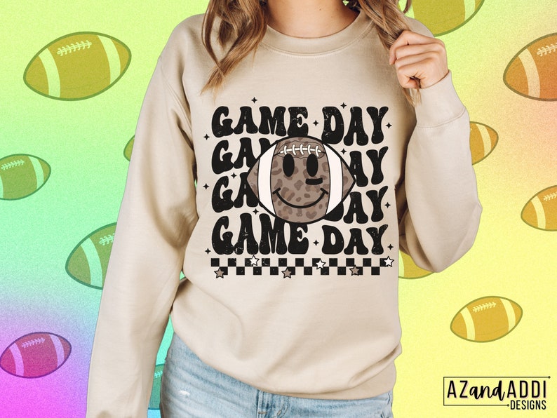 May include: A beige sweatshirt featuring the text "GAME DAY" repeated three times, with a football graphic in the middle. The football has a smiling face and a leopard print pattern. The sweatshirt has a crew neck and long sleeves.
