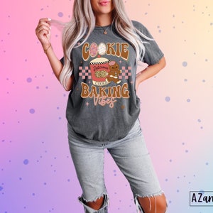 May include: Dark gray t-shirt with a graphic that says "Cookie Baking Vibes." The design includes a cookie, a gingerbread man, and a checkered pattern. The shirt is a relaxed fit and is paired with light-wash jeans.