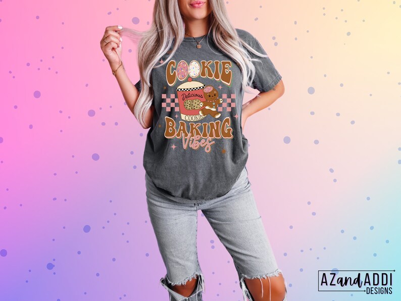 May include: Dark gray t-shirt with a graphic that says "Cookie Baking Vibes." The design includes a cookie, a gingerbread man, and a checkered pattern. The shirt is a relaxed fit and is paired with light-wash jeans.