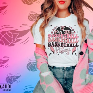 May include: White t-shirt with the words "BASKETBALL BASKETBALL BASKETBALL Vibes" in pink and black, featuring a basketball and lightning bolt design. The person is wearing a pink and green patterned cardigan.