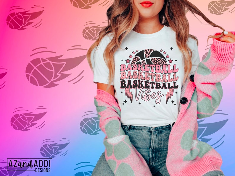 May include: White t-shirt with the words "BASKETBALL BASKETBALL BASKETBALL Vibes" in pink and black, featuring a basketball and lightning bolt design. The person is wearing a pink and green patterned cardigan.