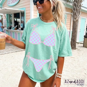 K&ouml;nnte beinhalten: Hellgr&uuml;nes T-Shirt mit einem rosa-blau karierten Bikini-Design. Das Oberteil und der Unterteil des Bikinis sind rosa umrandet und mit Schleifen versehen. Das Shirt ist mit "AZANDADDI DESIGNS" unten rechts versehen.