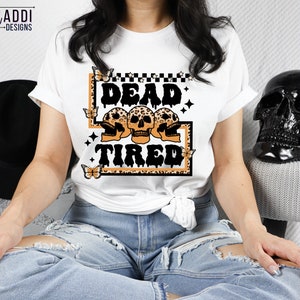 Op de afbeelding: Wit T-shirt met een afbeelding van drie schedels met de tekst "Dead & Tired" in zwarte en oranje letters. De afbeelding is omgeven door een luipaardprint rand en vlinders.