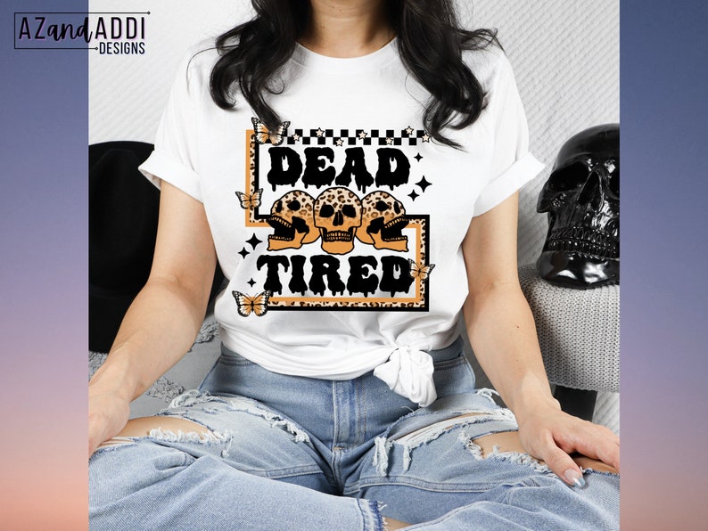 Op de afbeelding: Wit T-shirt met een afbeelding van drie schedels met de tekst "Dead & Tired" in zwarte en oranje letters. De afbeelding is omgeven door een luipaardprint rand en vlinders.