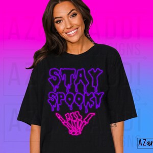 May include: Black t-shirt with a purple and pink neon design that says "Stay Spooky" with a skeleton hand giving the shaka sign.