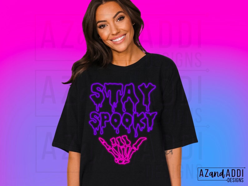 May include: Black t-shirt with a purple and pink neon design that says "Stay Spooky" with a skeleton hand giving the shaka sign.