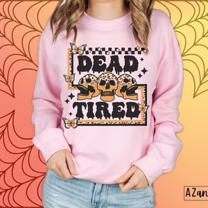 Op de afbeelding: Roze sweatshirt met een grafische print van drie schedels met luipaardprint en de tekst "Dead Tired" in zwarte druppelende letters. De grafiek is omgeven door een zwart-witte geruite rand en vlinders.