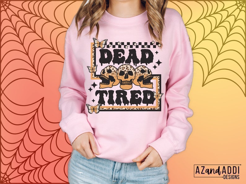 Op de afbeelding: Roze sweatshirt met een grafische print van drie schedels met luipaardprint en de tekst "Dead Tired" in zwarte druppelende letters. De grafiek is omgeven door een zwart-witte geruite rand en vlinders.