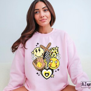 May include: A light pink sweatshirt featuring a softball-themed graphic. The design includes smiley faces, a baseball bat, a glove, a softball, and a heart. The graphic is in yellow, black, and white. The text "AZAND ADDI DESIGNS" is in the bottom right corner.