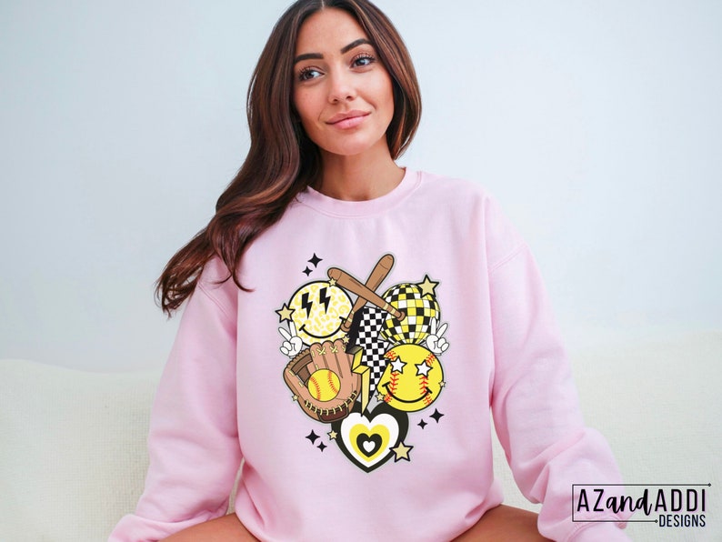 May include: A light pink sweatshirt featuring a softball-themed graphic. The design includes smiley faces, a baseball bat, a glove, a softball, and a heart. The graphic is in yellow, black, and white. The text "AZAND ADDI DESIGNS" is in the bottom right corner.