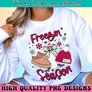 May include: White sweatshirt with a red, green and black graphic design that says "Freezin' Season". The design includes a snowflake, a red and white hat, a pair of brown boots and a green and white bow.
