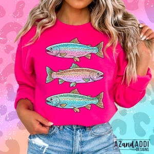 Puede incluir: Una sudadera rosa brillante con tres ilustraciones de peces coloridos. Los peces son de color azul, marr&oacute;n y rosa, con escamas y aletas detalladas. El dise&ntilde;o es un gr&aacute;fico l&uacute;dico y llamativo, perfecto para los amantes de la pesca o los temas acu&aacute;ticos.