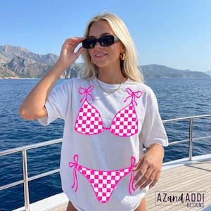 K&ouml;nnte beinhalten: Wei&szlig;es T-Shirt mit einem pink-wei&szlig; karierten Bikini-Design. Das Bikini-Oberteil und -Unterteil sind pink umrandet, mit pinken Schleifen. Das Shirt hat den Text "AZandADDI DESIGNS" unten rechts.