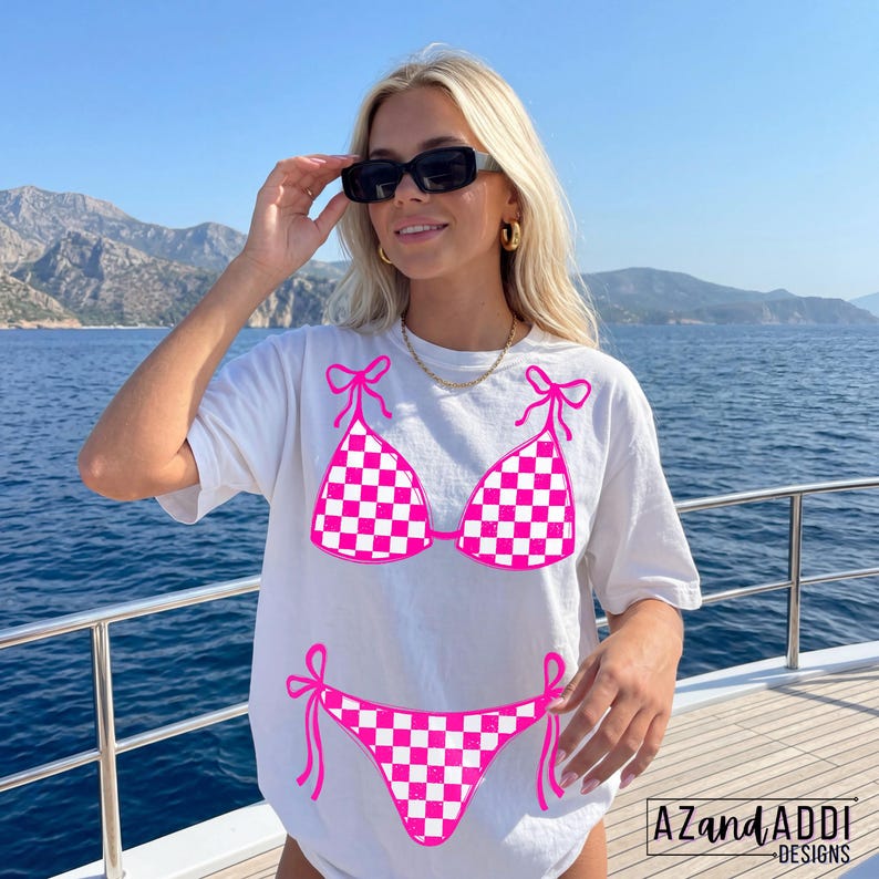 K&ouml;nnte beinhalten: Wei&szlig;es T-Shirt mit einem pink-wei&szlig; karierten Bikini-Design. Das Bikini-Oberteil und -Unterteil sind pink umrandet, mit pinken Schleifen. Das Shirt hat den Text "AZandADDI DESIGNS" unten rechts.