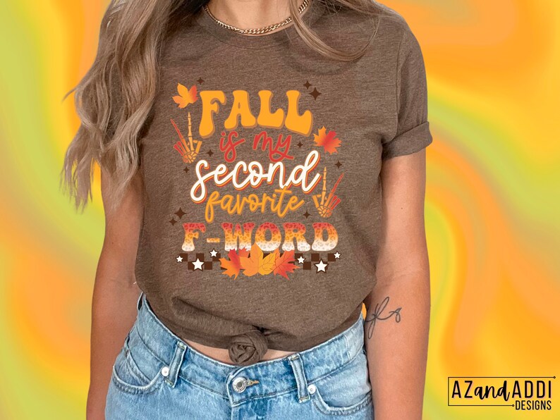 Pu&ograve; includere: T-shirt marrone con la scritta "FALL is my second favorite F-WORD" in arancione, bianco e marrone. Il design include foglie autunnali, stelle e una mano di scheletro. Abbigliamento casual a tema autunnale.
