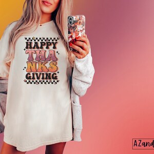 May include: A white t-shirt with a retro-style graphic that says "Happy Thanks Giving" in pink, orange, and yellow. The graphic has a checkered pattern and stars around it.