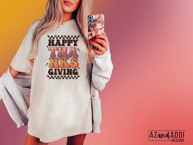 May include: A white t-shirt with a retro-style graphic that says "Happy Thanks Giving" in pink, orange, and yellow. The graphic has a checkered pattern and stars around it.