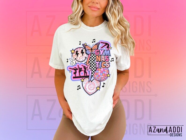 May include: White t-shirt with a graphic design featuring a pink and purple butterfly, a disco ball, a heart with a tie-dye pattern, and the text "Gymnastics".