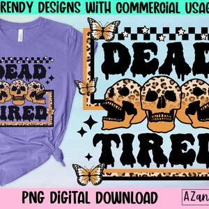 May include: A purple t-shirt with a graphic design of three skulls with leopard print and the text "Dead Tired" in black dripping letters. The design is surrounded by a black and white checkered border with white stars.