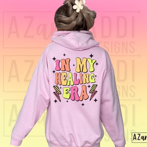 K&ouml;nnte beinhalten: Rosafarbener Hoodie mit dem Text "In My Healing Era" in bunten Retro-Lettern mit Blitzen und Sternen.