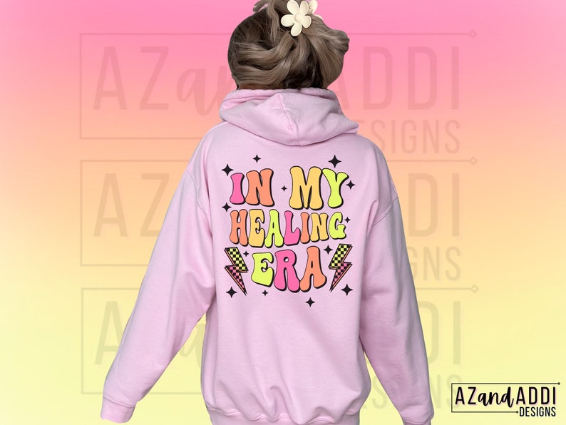 K&ouml;nnte beinhalten: Rosafarbener Hoodie mit dem Text "In My Healing Era" in bunten Retro-Lettern mit Blitzen und Sternen.