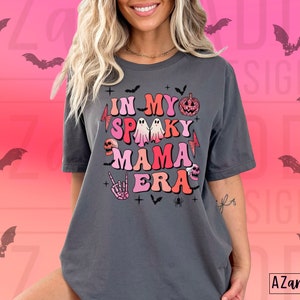 May include: Dark gray t-shirt with the text "IN MY SPOOKY MAMA ERA" in colorful, retro-style lettering. The design includes Halloween-themed graphics like a pumpkin, ghost, skull, and bats. The shirt has a relaxed fit and short sleeves.