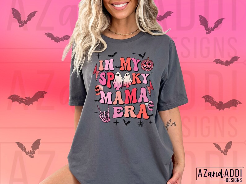 May include: Dark gray t-shirt with the text "IN MY SPOOKY MAMA ERA" in colorful, retro-style lettering. The design includes Halloween-themed graphics like a pumpkin, ghost, skull, and bats. The shirt has a relaxed fit and short sleeves.