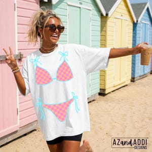 K&ouml;nnte beinhalten: Wei&szlig;es T-Shirt mit einem pink-orange karierten Bikini-Design und hellblauen Akzenten. Das l&auml;ssige T-Shirt hat eine lockere Passform. Das Bild ist vor bunten Strandh&uuml;tten aufgenommen, was auf ein Sommerthema hindeutet.
