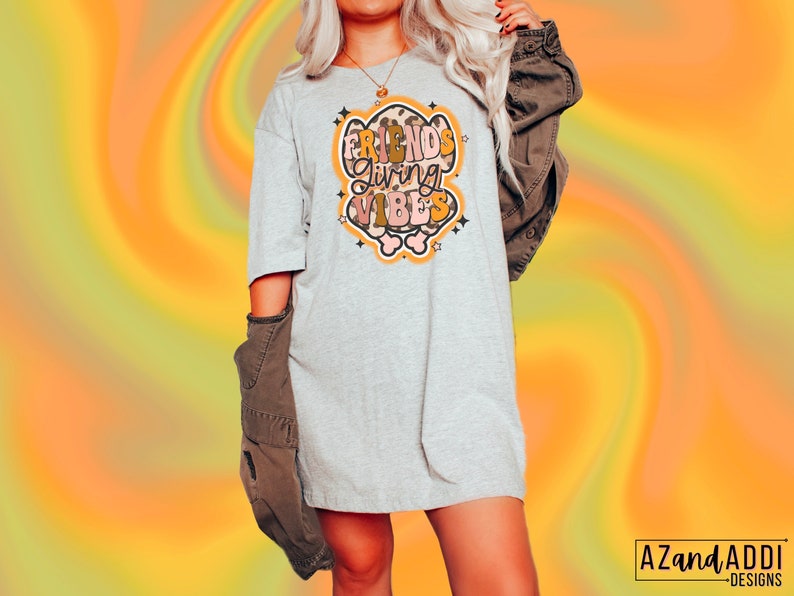 May include: A light gray t-shirt dress with the words "FRIENDS GIVING VIBES" in a retro design. The graphic is in shades of brown, orange, and pink, with star accents. The dress is paired with a brown jacket. The background is a swirl of orange and yellow.